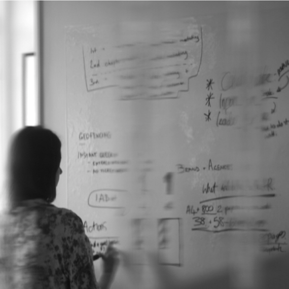 Greyscale photo of person writing on a whiteboard in an office
